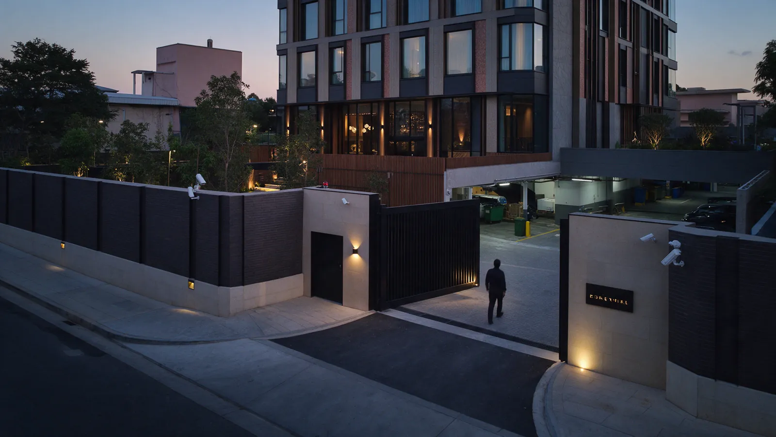 Illustrative hotel service perimeter with a distant person crossing an open gate at dusk.